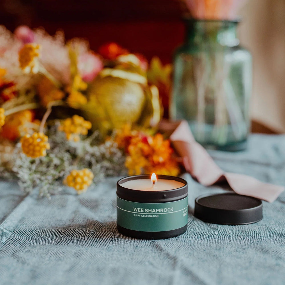 Candle labeled 'Wee Shamrock' on a table with flowers and a jar in the background